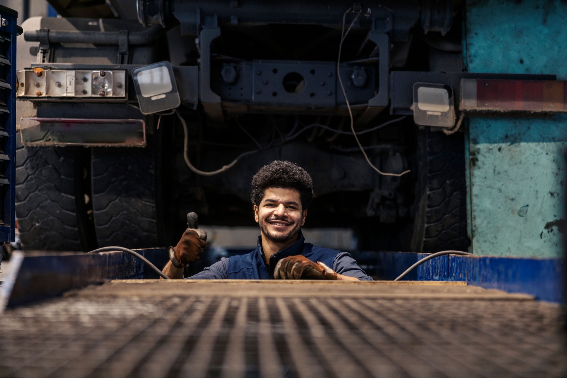 Portrait of smiling interracial technician in mechanic pit giving thumbs up and repairing big truck.